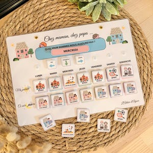 Peut inclure: Un tableau magnétique coloré avec un emploi du temps quotidien pour les enfants. Le tableau présente une illustration de maison, les mots "Chez maman, chez papa..." et "Quel jour sommes nous aujourd'hui?"  Le tableau comprend les jours de la semaine, des activités comme "école", "sport", et "centre de loisirs", et des lieux comme "chez maman" et "chez papa".