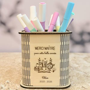 Peut inclure: Un pot à crayons en bois avec un design décoratif et le texte "MERCI MAÎTRE pour cette belle année". Le pot contient des marqueurs colorés. Le nom "Chloé" et l'année "2025-2026" sont également visibles.