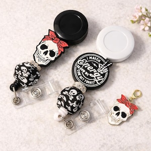 Puede incluir: Una colección de portatarjetas retráctiles y un dije con diseños de calaveras. Los portatarjetas tienen cuentas de calaveras blancas y negras, y una calavera con un lazo rojo. Un portatarjetas tiene el texto "Match Energy".
