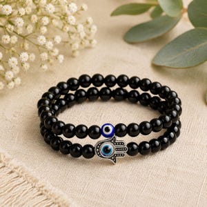 Puede incluir: Un conjunto de dos pulseras elásticas hechas de cuentas negras lisas y redondas. Una pulsera presenta una pequeña cuenta redonda de ojo turco azul y blanco. La segunda pulsera incluye un dije de mano de Hamsa plateado con un ojo turco en el centro.