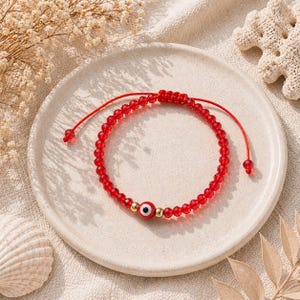May include: Red beaded bracelet with an evil eye charm. The bracelet features small, faceted red beads strung on a red cord. A gold bead with a red and white evil eye charm is centered on the bracelet. The bracelet is displayed on a white plate.
