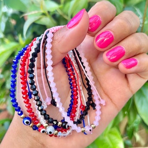 May include: A collection of beaded bracelets in various colors, including blue, red, black, and pink. Some bracelets feature gold accents and evil eye charms. The bracelets are strung on cords and are displayed against a blurred green background.