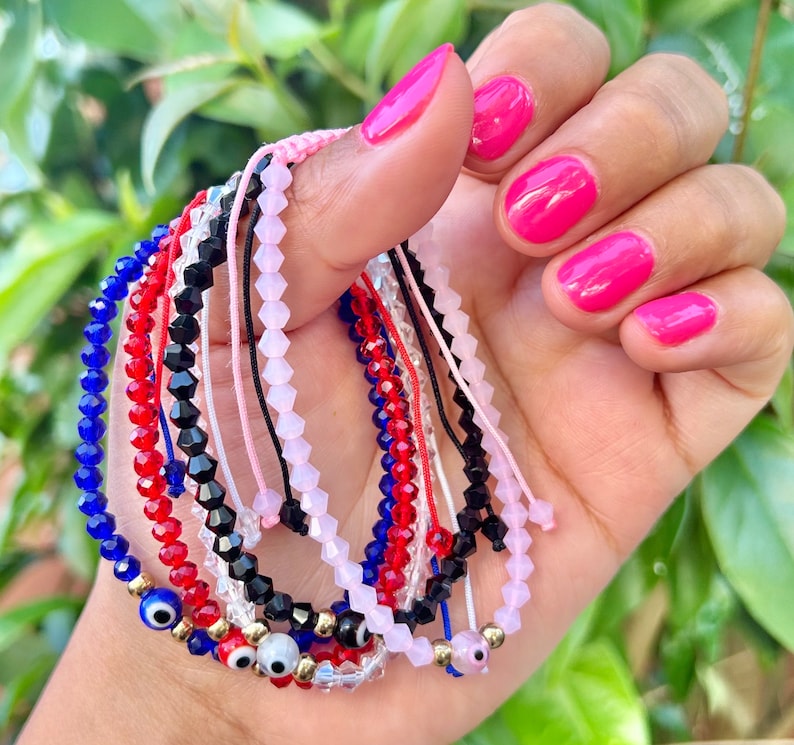 May include: A collection of beaded bracelets in various colors, including blue, red, black, and pink. Some bracelets feature gold accents and evil eye charms. The bracelets are strung on cords and are displayed against a blurred green background.