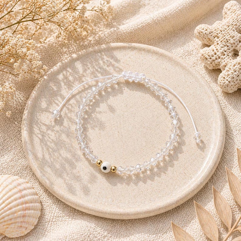 May include: A clear crystal beaded bracelet with a white cord and a small white bead with a black dot and gold beads. The bracelet is on a beige plate with a seashell and dried flowers.