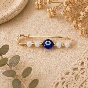 Puede incluir: Alfiler de seguridad dorado con una cuenta de ojo turco azul y cuentas blancas. El alfiler est&aacute; adornado con una cuenta central azul oscuro con un dise&ntilde;o de ojo blanco y negro, flanqueada por cuentas blancas y doradas.