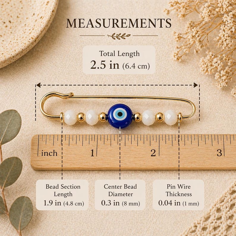 Puede incluir: Alfiler de seguridad dorado con un amuleto de ojo turco. El alfiler presenta cuentas blancas y doradas, y una cuenta de ojo turco azul y blanca. Longitud total: 6,4 cm.