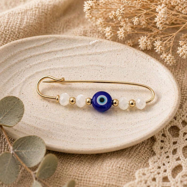 Puede incluir: Alfiler de seguridad dorado con una cuenta azul de ojo turco, cuentas blancas y detalles dorados. El alfiler se muestra en un plato color crema, mostrando un dise&ntilde;o de joyer&iacute;a &uacute;nico. Este accesorio es perfecto para a&ntilde;adir un toque de estilo.