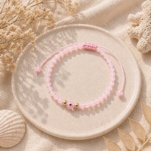 May include: A pink beaded bracelet with an evil eye charm, displayed on a beige plate. The bracelet features small, round pink beads and gold-colored accents. The evil eye charm is pink with a blue center. The bracelet is adjustable with a sliding knot.