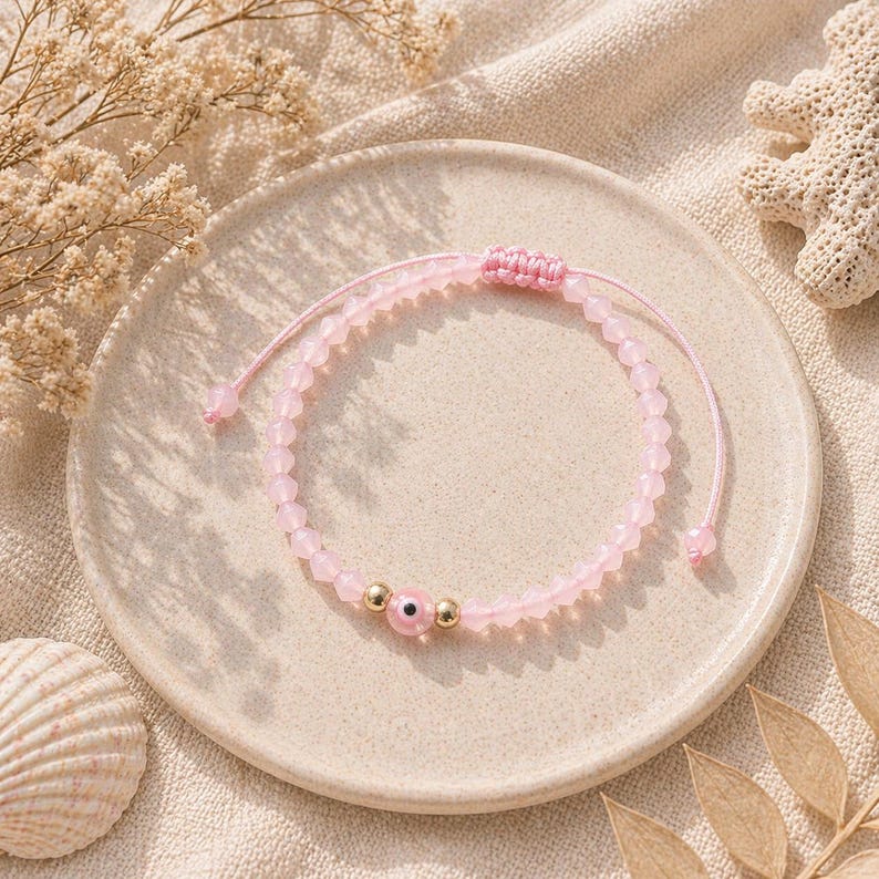 May include: A pink beaded bracelet with an evil eye charm, displayed on a beige plate. The bracelet features small, round pink beads and gold-colored accents. The evil eye charm is pink with a blue center. The bracelet is adjustable with a sliding knot.