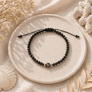 May include: Black beaded bracelet with an evil eye charm, set on a neutral-toned plate. The bracelet features small, faceted black beads and a central charm with a blue and white eye design, framed by gold-colored beads.