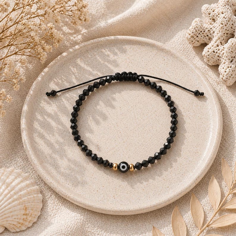 May include: Black beaded bracelet with an evil eye charm, set on a neutral-toned plate. The bracelet features small, faceted black beads and a central charm with a blue and white eye design, framed by gold-colored beads.