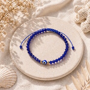 May include: A blue beaded bracelet with an evil eye charm. The bracelet features small, faceted blue beads and a gold-colored bead with a blue and white evil eye design. The bracelet is on a neutral-colored surface.