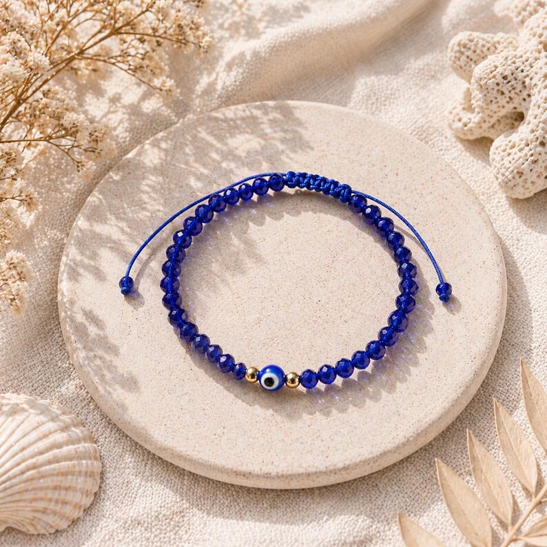 May include: A blue beaded bracelet with an evil eye charm. The bracelet features small, faceted blue beads and a gold-colored bead with a blue and white evil eye design. The bracelet is on a neutral-colored surface.