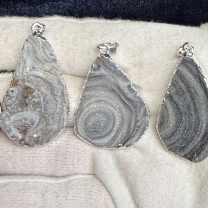 May include: Three silver pendant charms with a teardrop shape. Each pendant is made of a light gray agate with a swirling pattern. The agate is set in a silver bezel.