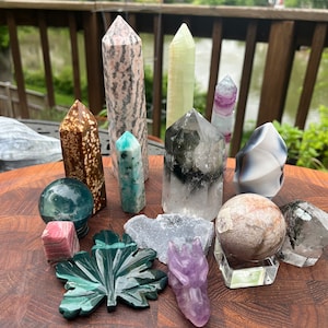 May include: A collection of polished and carved crystals and stones, including a green leaf, a dragon, a sphere, and various points. The crystals are arranged on a wooden surface.