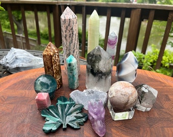 Peut inclure: Une collection de cristaux et de pierres polies et sculptées, comprenant une feuille verte, un dragon, une sphère et divers pointes. Les cristaux sont disposés sur une surface en bois.