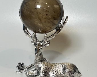 May include: A smoky quartz crystal sphere sits atop a silver deer figurine. The deer is lying down with its head tilted up, holding the sphere in its antlers. The deer has a flower on its tail.