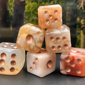 May include: Six agate dice with copper accents. The dice are various shades of brown, white, and pink. The dice are arranged in a pyramid shape.