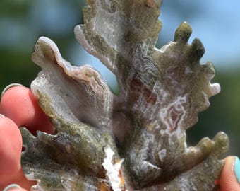 May include: A carved stone leaf sculpture with a natural, organic appearance. The leaf features a mix of green, white, and brown tones, with intricate veining and a textured surface. The sculpture is held against a blurred background.