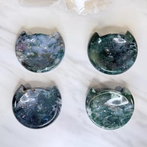 May include: Four polished moss agate stones carved into circular shapes with two small pointed ears at the top. Each stone features unique natural patterns of deep green, translucent white, and grey inclusions with a smooth, reflective surface.
