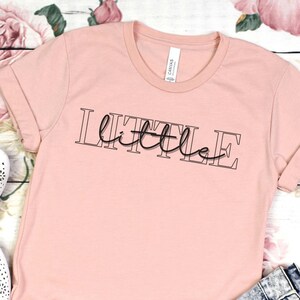 Puede incluir: Una camiseta rosa con el texto "LITTLE little" en letras negras. La camiseta está sobre una superficie de madera blanca con un par de zapatos blancos y unos pantalones cortos de mezclilla.
