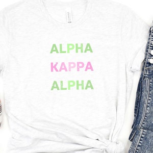 Könnte beinhalten: Ein weißes T-Shirt mit dem Aufdruck "ALPHA KAPPA ALPHA" in grünen und rosa Buchstaben.