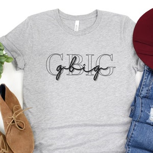 Puede incluir: Una camiseta gris con el texto "Go Big" en letras negras. El texto está contorneado en gris. La camiseta se muestra sobre un fondo blanco con un par de botas marrones, un par de pantalones vaqueros azules y un sombrero burdeos.