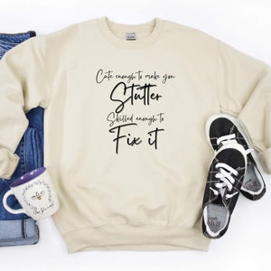 May include: A beige sweatshirt with the text "Cute enough to make you stutter. Skilled enough to fix it." printed in black.