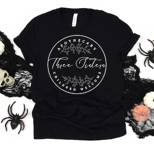 Pode incluir: Camiseta preta com um gráfico circular branco que diz "Apothecary Three Sisters Children Welcome". O gráfico apresenta um design floral.