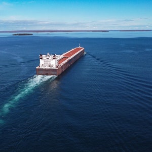 Może przedstawiać: Widok z lotu ptaka na duży statek towarowy płynący po głębokim, niebieskim morzu pod czystym niebem. Statek pozostawia biały ślad, a powierzchnia wody pokazuje jego ślad. W oddali widoczne są wyspy.