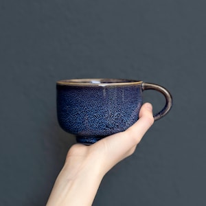 Puede incluir: Una taza de cerámica con un esmalte moteado en azul oscuro y negro. La taza presenta un cuerpo ancho y redondeado, una base corta y un asa curva en el lado derecho. La textura de la superficie parece granulada con tonos azules profundos.