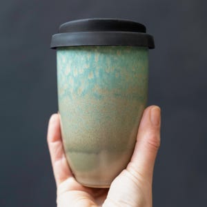 May include: A ceramic travel mug with a black lid. The mug has a speckled turquoise and tan glaze, transitioning to a darker green at the base. The mug is held against a dark background.