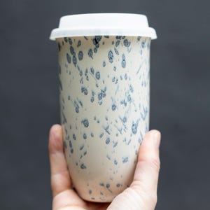 Puede incluir: Un termo de viaje de cerámica blanca con tapa blanca. La taza está decorada con patrones moteados azules. La taza se sostiene en una mano sobre un fondo gris oscuro. El termo de viaje está diseñado para mantener las bebidas calientes o frías.
