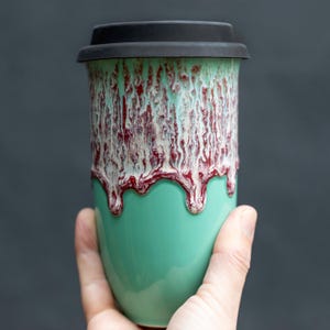Puede incluir: Una taza de viaje de cerámica de color turquesa con una tapa negra. La taza tiene un diseño marmoleado rojo y blanco.