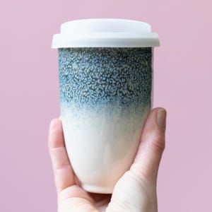 May include: A ceramic travel mug with a white lid. The mug has a speckled blue and white glaze that fades from blue at the top to white at the bottom. The mug is held in a hand against a pink background.