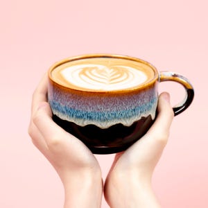May include: A ceramic coffee mug with a brown, blue, and black glaze, filled with a latte featuring a heart-shaped design in the foam. The mug is held by two hands against a pink background.