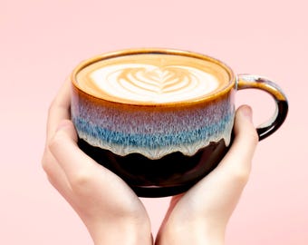 May include: A ceramic coffee mug with a brown, blue, and black glaze, filled with a latte featuring a heart-shaped design in the foam. The mug is held by two hands against a pink background.