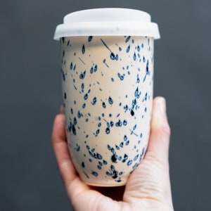 May include: A white ceramic travel mug with a blue and white speckled pattern. The mug has a white lid.
