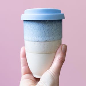 May include: A ceramic travel mug with a light blue silicone lid. The mug has a layered design with shades of blue and cream. The mug is held in a hand against a pink background. The mug is approximately 5 inches tall.