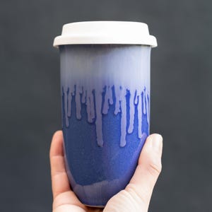 May include: A blue ceramic travel mug with a white lid. The mug has a gradient effect, transitioning from a lighter blue at the top to a darker blue at the bottom, with white drip-like accents. The mug is held in a hand.