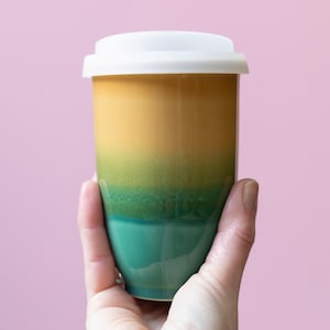 May include: A ceramic travel mug with a white lid. The mug features a gradient of colors, transitioning from yellow to green to teal. The mug is held in a hand against a pink background.