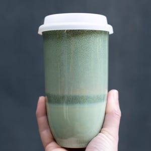 May include: A green ceramic travel mug with a white lid. The mug has a speckled, glazed finish and a cylindrical shape. The color gradient transitions from a darker green at the base to a lighter green at the top.