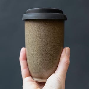 May include: A brown travel mug with a textured surface and a black lid. The reusable coffee cup is held in a hand against a dark background. The mug is designed for hot or cold beverages.