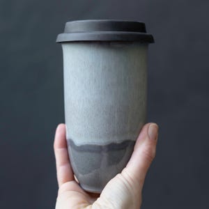 Może przedstawiać: Ceramiczny kubek podróżny o gładkim, zwężającym się cylindrycznym korpusie i matowym wykończeniu. Kubek posiada gradientową kolorystykę, przechodzącą od jasnej, bladej szarości u góry do głębokiej, ciemnej szarości u podstawy.