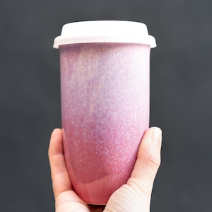 May include: A ceramic travel mug with a white lid. The mug has a gradient colour scheme, transitioning from a deep red at the base to a lighter pink near the top. The mug is held against a dark grey background.