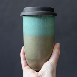 May include: A ceramic travel mug with a black lid. The mug is cylindrical with a two-tone glaze: a teal upper section and a tan lower section. The mug is held by a hand against a dark background.