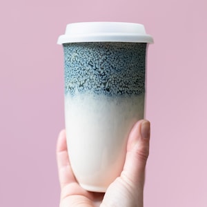 May include: A ceramic travel mug with a white lid. The mug has a white base that fades into a speckled blue-green pattern at the top. The mug is held against a pink background.