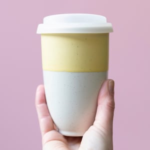 May include: A two-tone ceramic travel mug with a white lid. The top half is yellow, and the bottom half is white with small speckled details. The mug is held in a hand against a pink background. The mug is designed for hot or cold beverages.