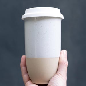 May include: A white and beige travel mug with a white lid. The upper portion of the mug is white with small black speckles, while the bottom is beige. The mug is held in a hand against a dark background.
