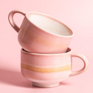 Könnte beinhalten: Zwei rosa Keramikbecher, die übereinander gestapelt sind. Jeder Becher hat einen Henkel und einen weißen Innenraum. Ein gelb-oranges Band verläuft um die Mitte des unteren Bechers. Der Hintergrund ist einfarbig rosa.